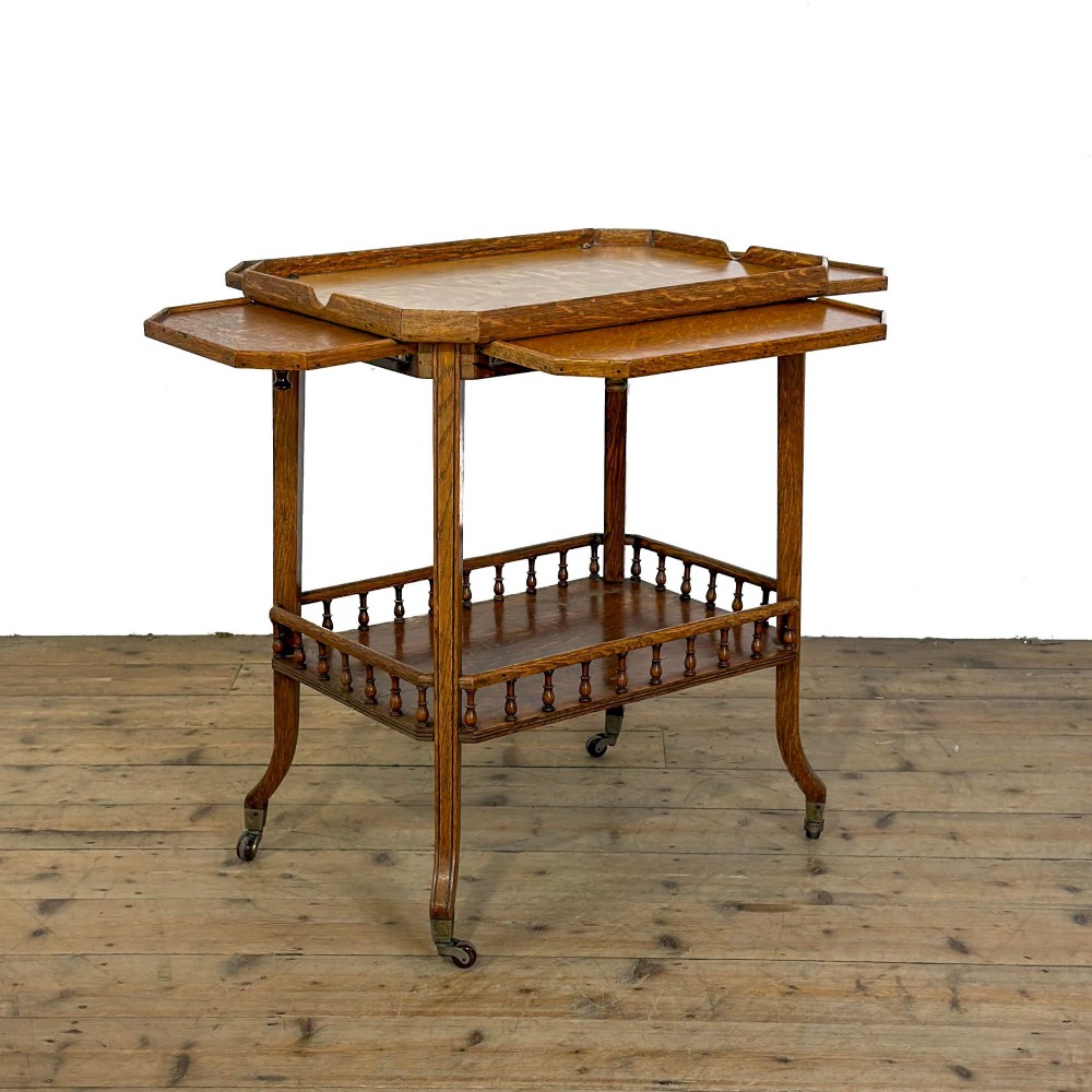 An Edwardian medium oak two-tier tea trolley featuring drop-leaf side panels, a spindle-turned gallery on the lower shelf, and brass castors. View VI