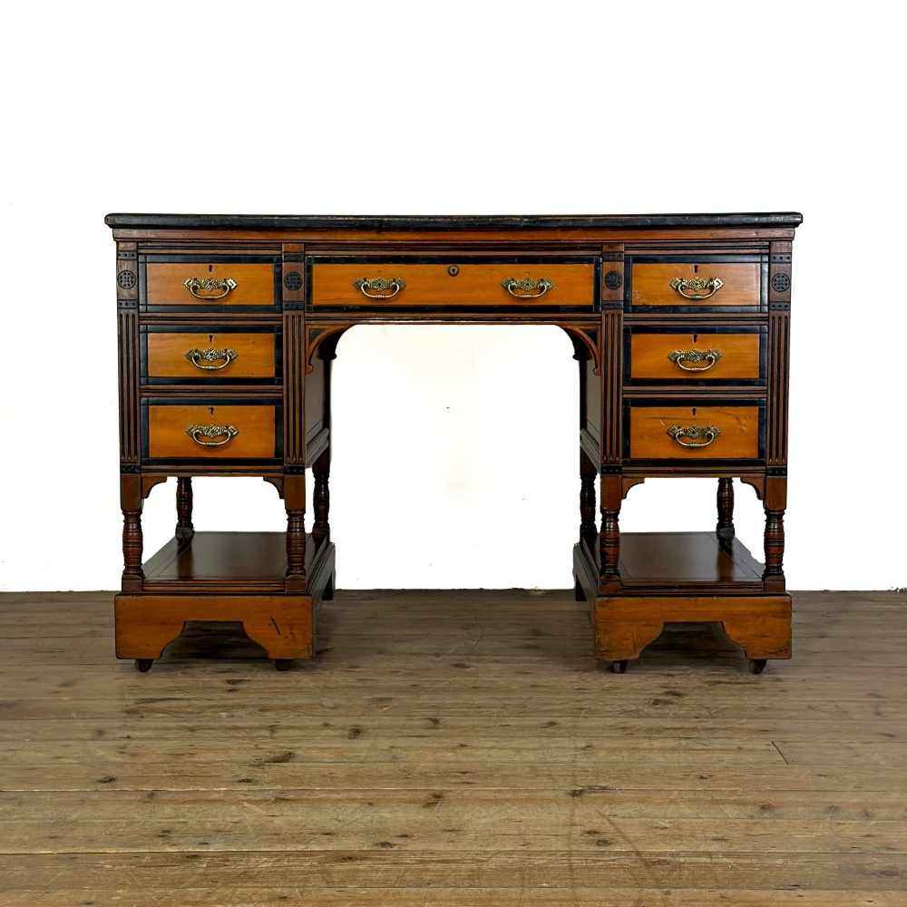 An Aesthetic Movement double-sided partners desk crafted from warm brown wood with black ebonised mouldings, brass drop handles, central kneehole drawer, twin pedestals, open lower shelves, and turned columns.