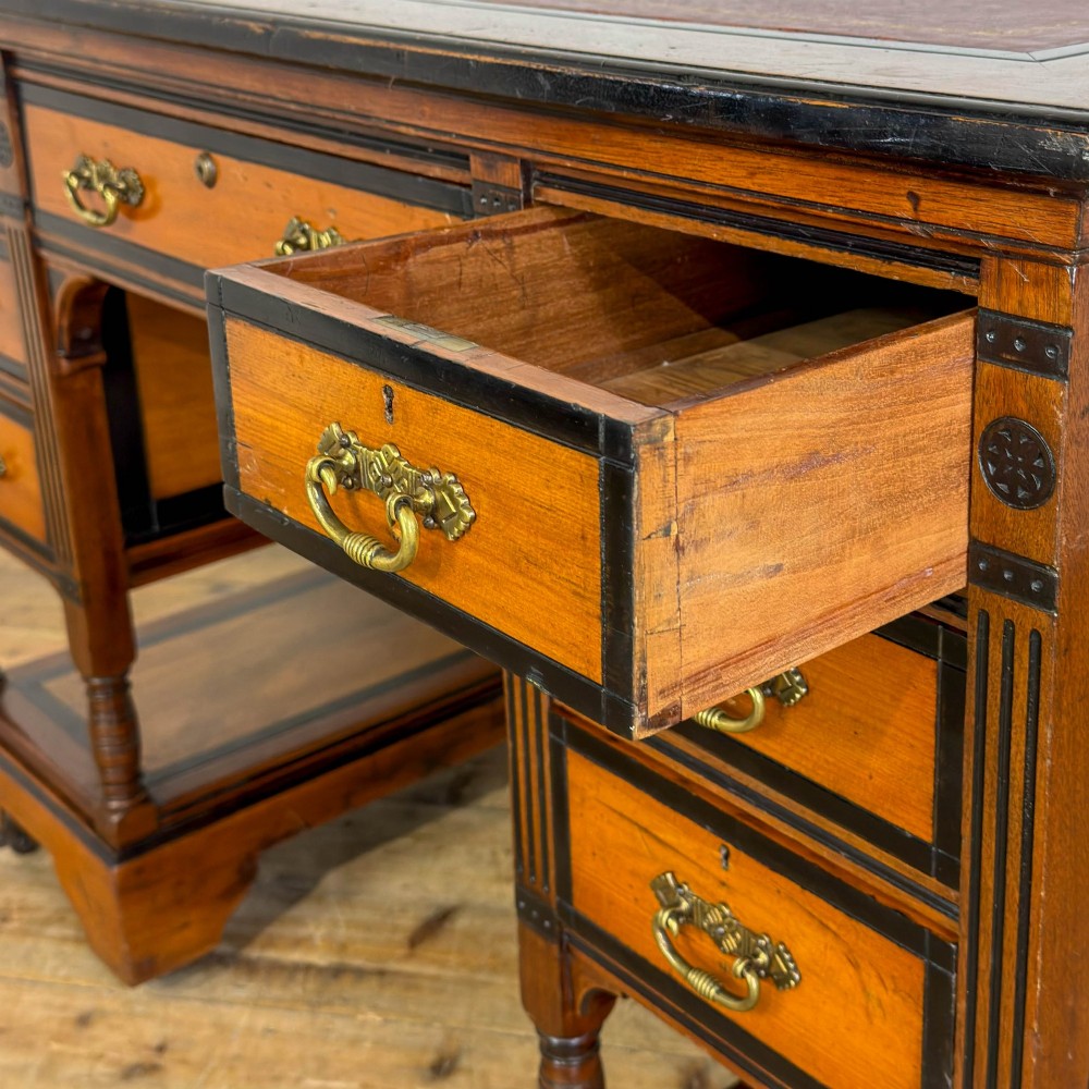 An Aesthetic Movement double-sided partners desk crafted from warm brown wood with black ebonised mouldings, brass drop handles, central kneehole drawer, twin pedestals, open lower shelves, and turned columns. View IV