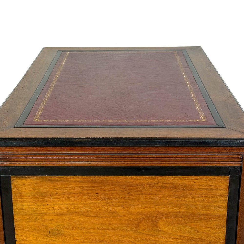 An Aesthetic Movement double-sided partners desk crafted from warm brown wood with black ebonised mouldings, brass drop handles, central kneehole drawer, twin pedestals, open lower shelves, and turned columns. View VI