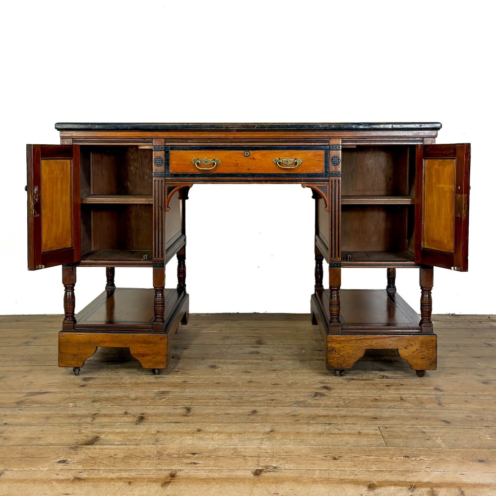 An Aesthetic Movement double-sided partners desk crafted from warm brown wood with black ebonised mouldings, brass drop handles, central kneehole drawer, twin pedestals, open lower shelves, and turned columns. View VII