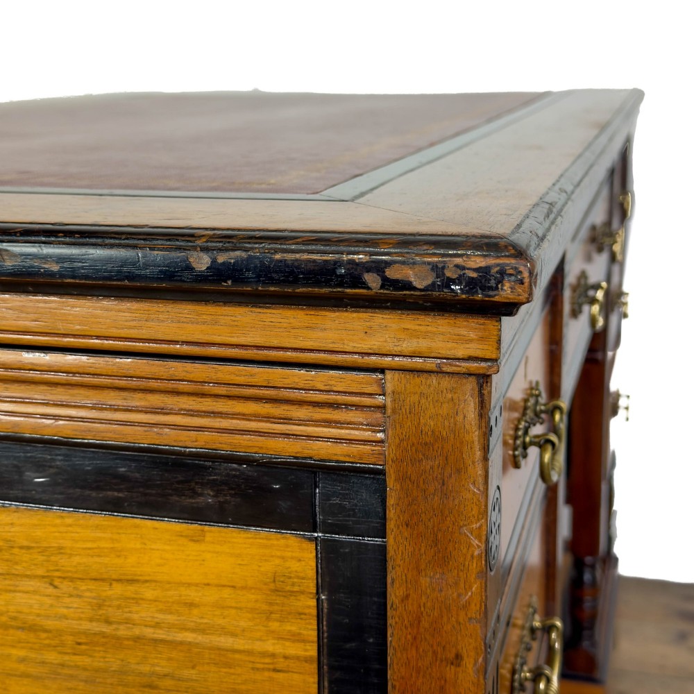 An Aesthetic Movement double-sided partners desk crafted from warm brown wood with black ebonised mouldings, brass drop handles, central kneehole drawer, twin pedestals, open lower shelves, and turned columns. View X