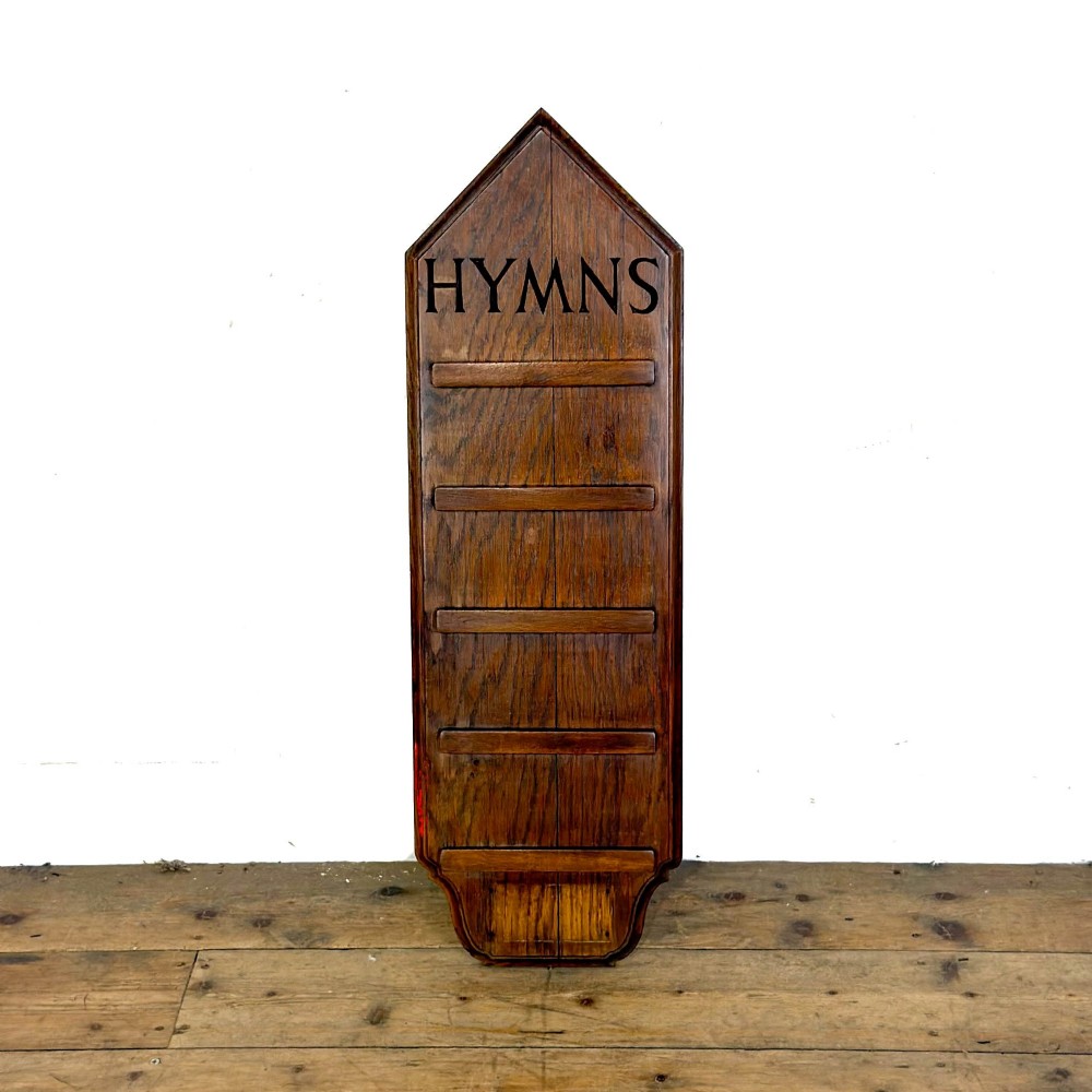 An antique solid oak hymn board featuring a pointed arched top with the word HYMNS cut out above five horizontal display slots.