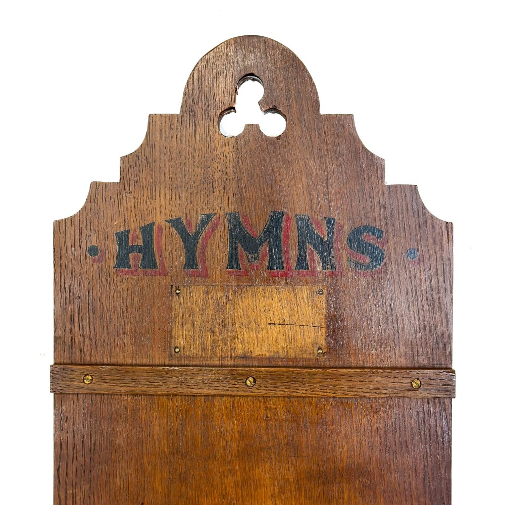 An antique dark oak church hymn board featuring a trefoil cutout at the carved top, painted text reading HYMNS, multiple horizontal slots, and an accompanying wooden box. View VIII