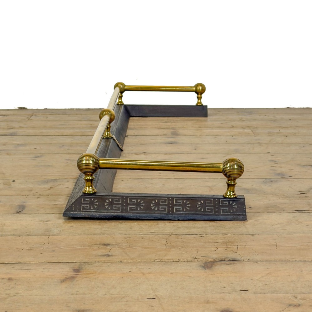 An antique Victorian fireplace fender featuring a dark metal base with stamped geometric patterns and an elevated brass rail supported by ribbed finials.