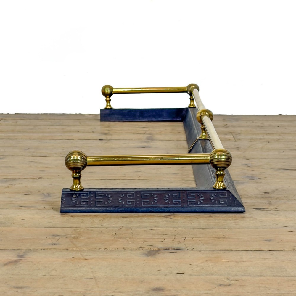 An antique Victorian fireplace fender featuring a dark metal base with stamped geometric patterns and an elevated brass rail supported by ribbed finials. View V
