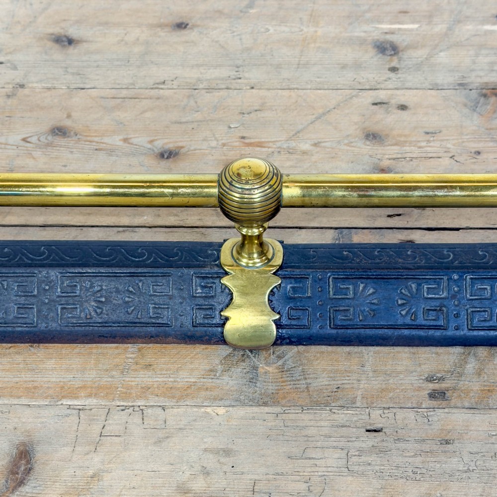 An antique Victorian fireplace fender featuring a dark metal base with stamped geometric patterns and an elevated brass rail supported by ribbed finials. View VI