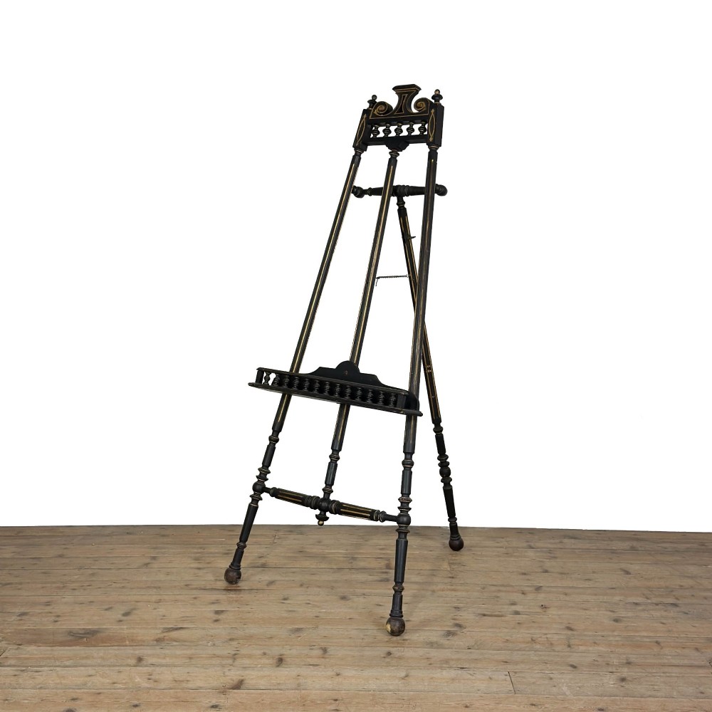 An ebonised wooden floor-standing easel with turned legs, geometric carved details, and gold leaf highlights set against a plain background.
