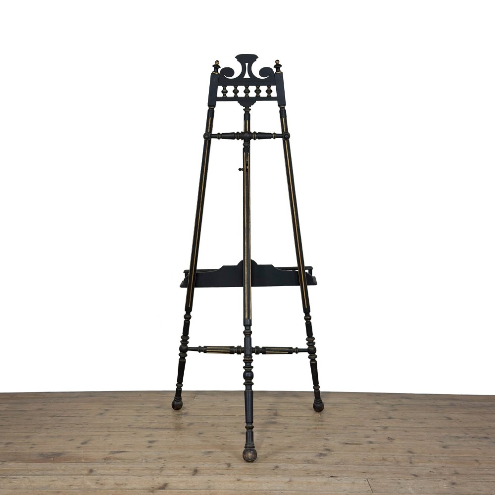 An ebonised wooden floor-standing easel with turned legs, geometric carved details, and gold leaf highlights set against a plain background. View II