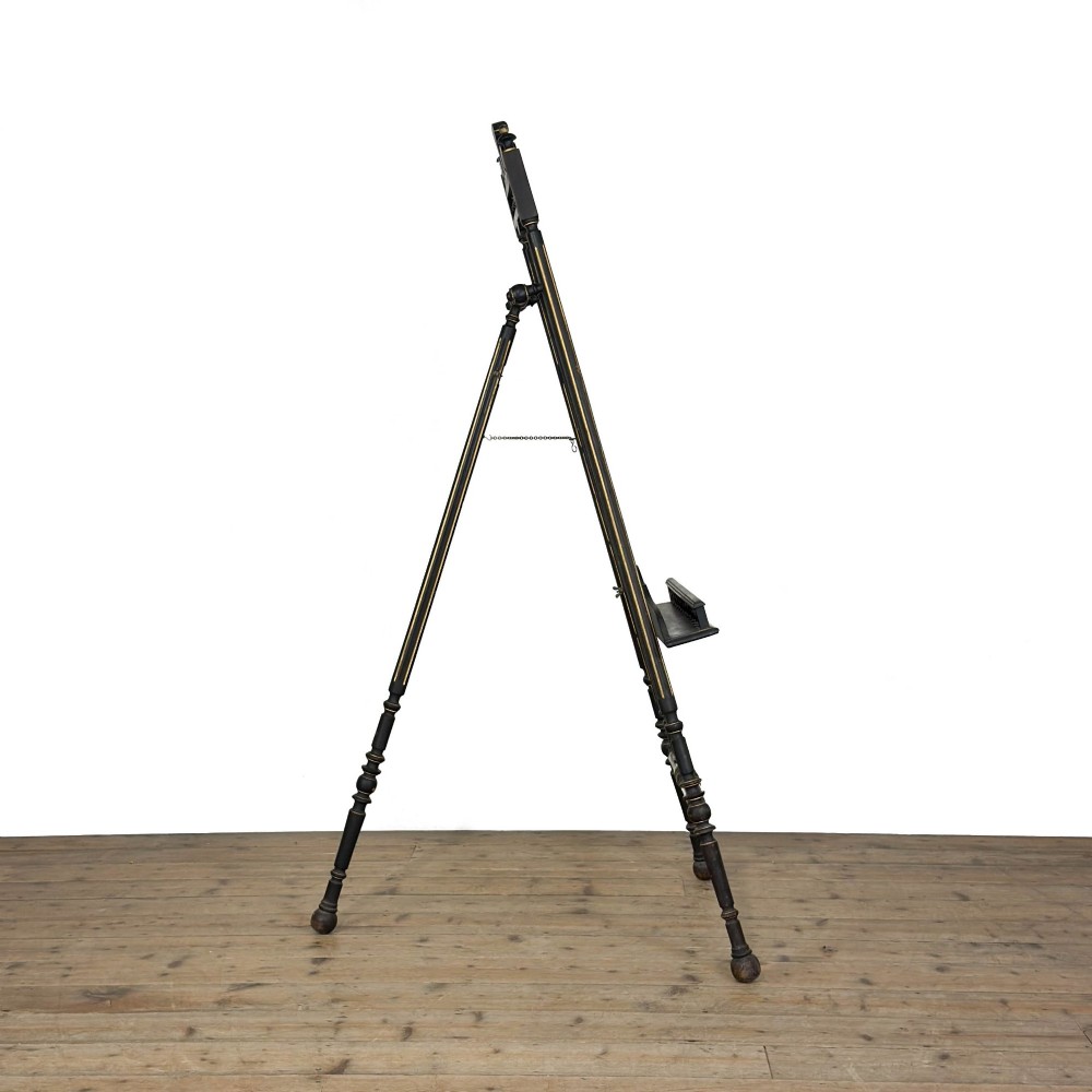 An ebonised wooden floor-standing easel with turned legs, geometric carved details, and gold leaf highlights set against a plain background. View III