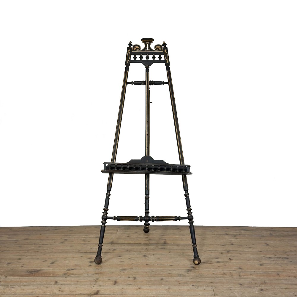 An ebonised wooden floor-standing easel with turned legs, geometric carved details, and gold leaf highlights set against a plain background. View IV