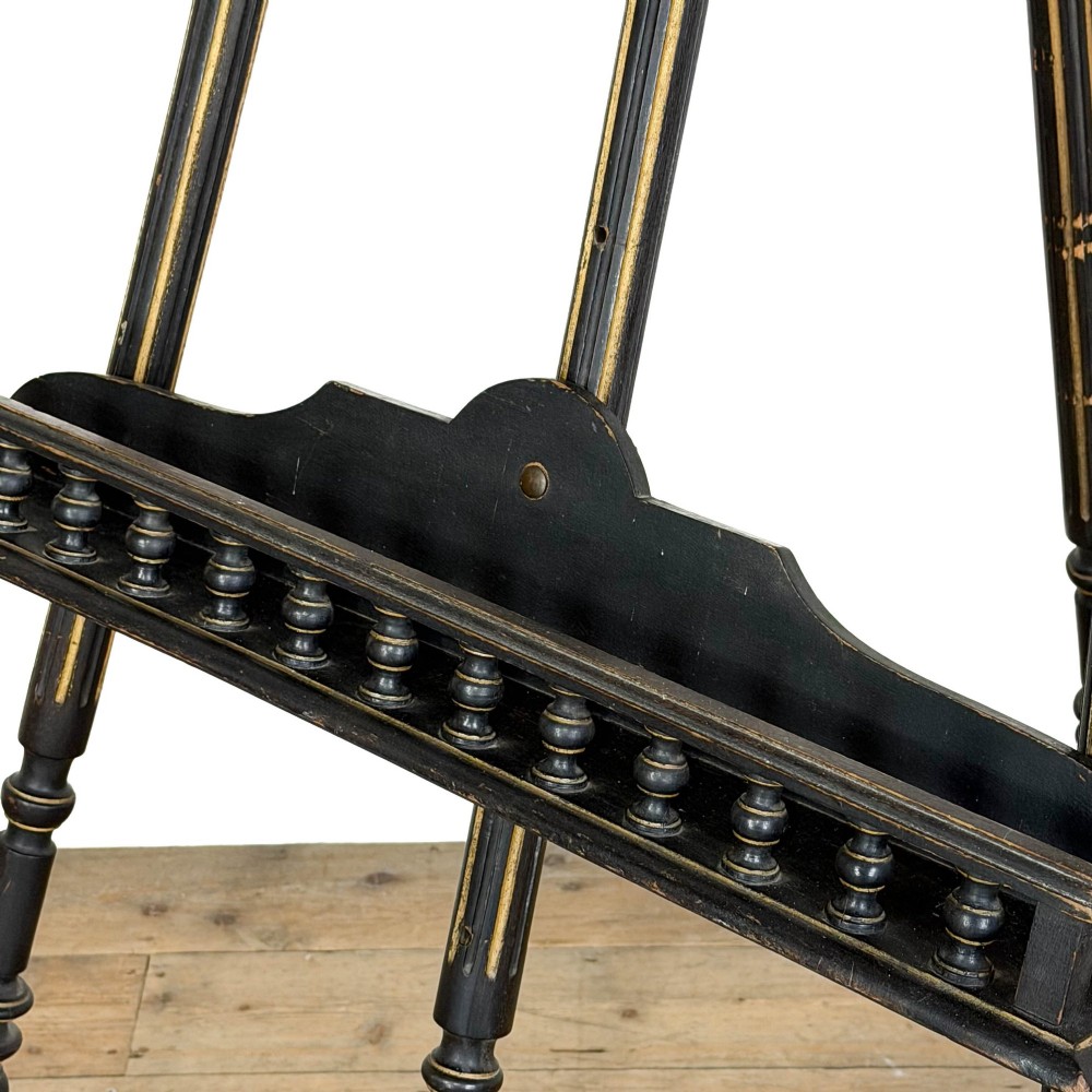 An ebonised wooden floor-standing easel with turned legs, geometric carved details, and gold leaf highlights set against a plain background. View V
