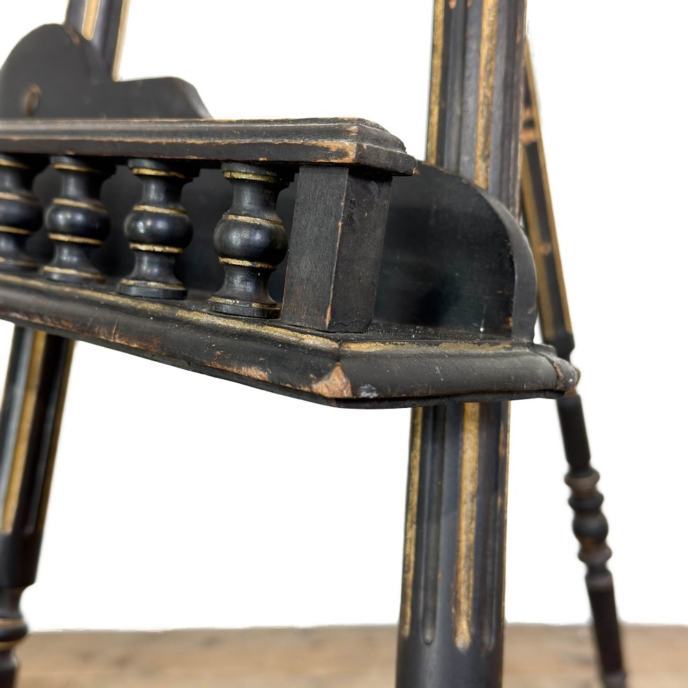 An ebonised wooden floor-standing easel with turned legs, geometric carved details, and gold leaf highlights set against a plain background. View VI