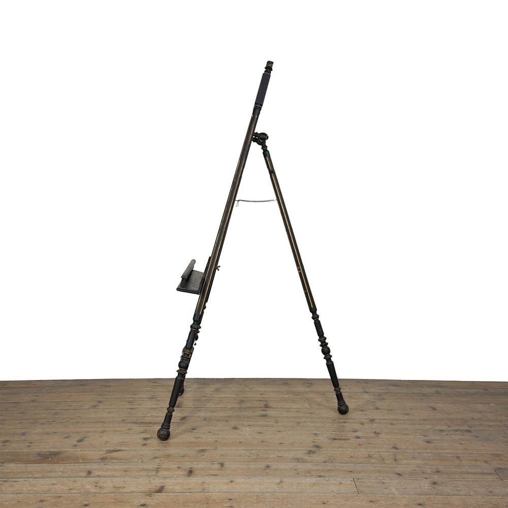 An ebonised wooden floor-standing easel with turned legs, geometric carved details, and gold leaf highlights set against a plain background. View VII