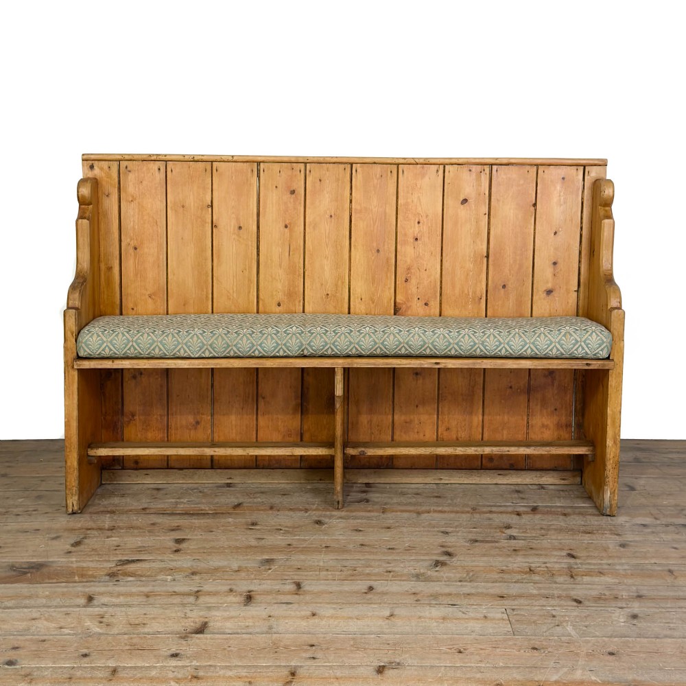 An antique pine church pew bench featuring a high vertical plank back, shaped scrolled side arms, a lower foot rail, and a patterned green fabric seat cushion.