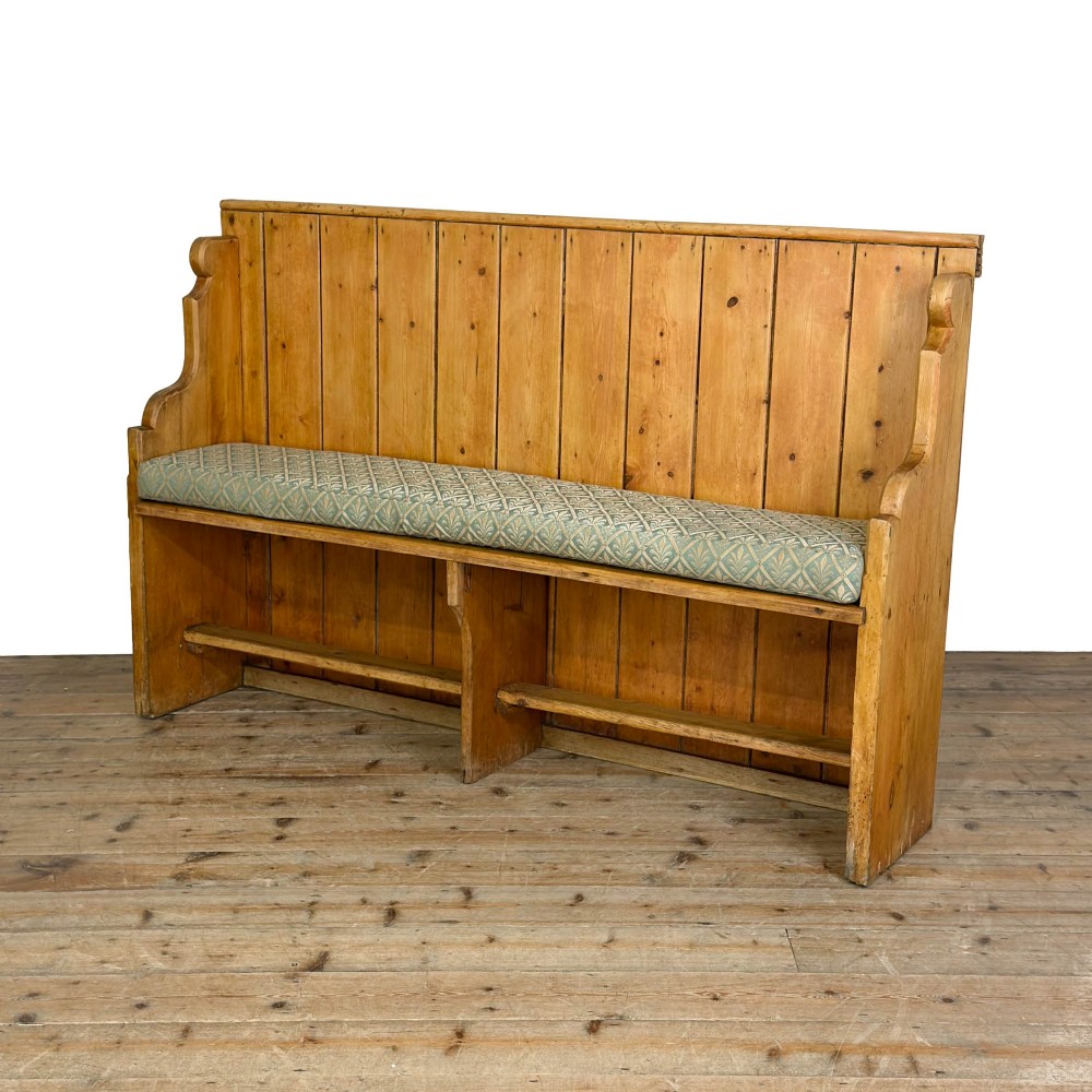 An antique pine church pew bench featuring a high vertical plank back, shaped scrolled side arms, a lower foot rail, and a patterned green fabric seat cushion. View III