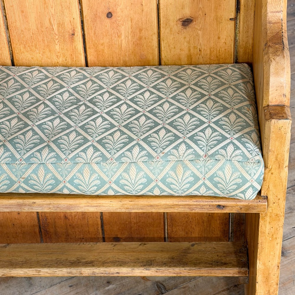 An antique pine church pew bench featuring a high vertical plank back, shaped scrolled side arms, a lower foot rail, and a patterned green fabric seat cushion. View V