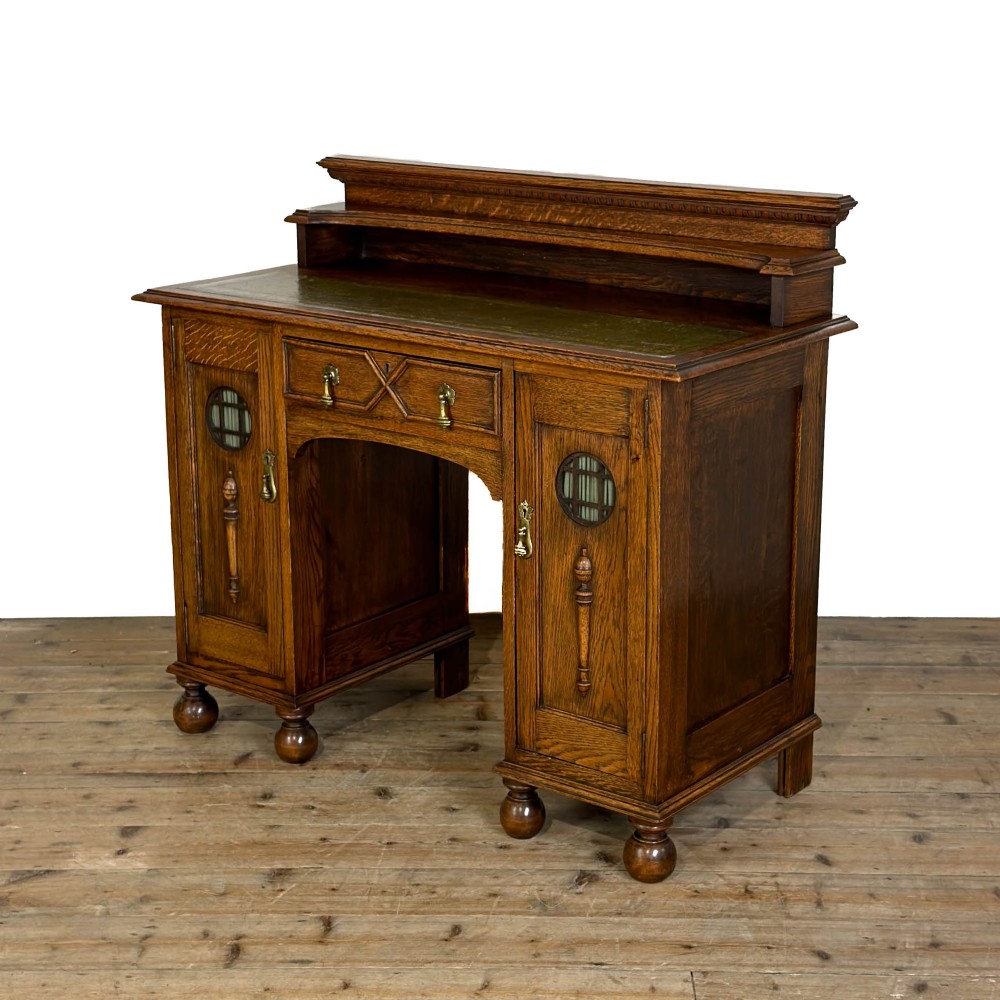 An Arts and Crafts solid oak kneehole desk featuring a raised top gallery shelf, central drawer with geometric moulding, drop handles, and side cupboards with circular gridded glass panels. View II