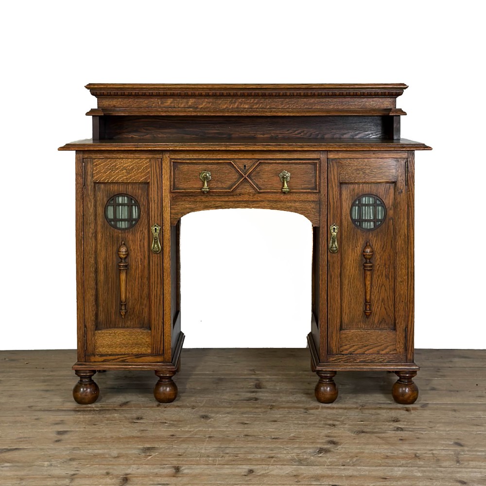 An Arts and Crafts solid oak kneehole desk featuring a raised top gallery shelf, central drawer with geometric moulding, drop handles, and side cupboards with circular gridded glass panels.