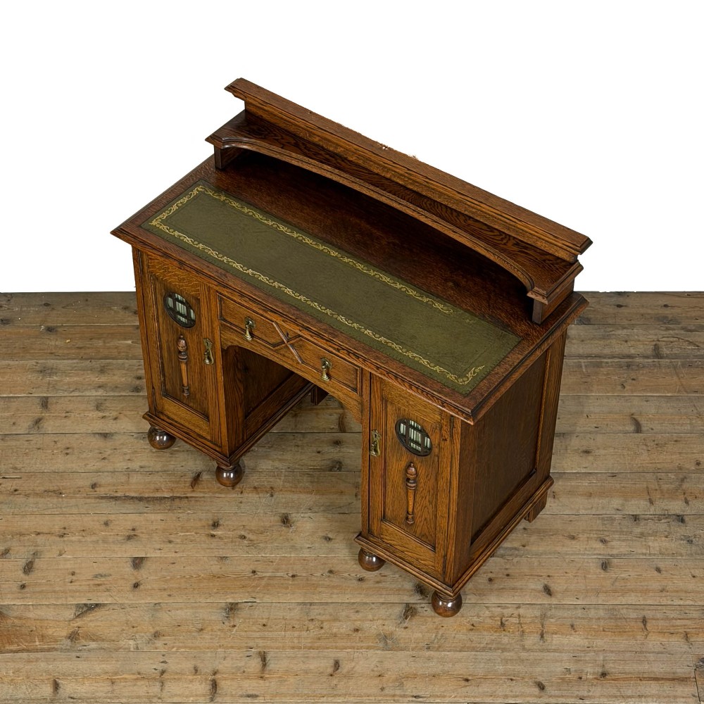 An Arts and Crafts solid oak kneehole desk featuring a raised top gallery shelf, central drawer with geometric moulding, drop handles, and side cupboards with circular gridded glass panels. View IV