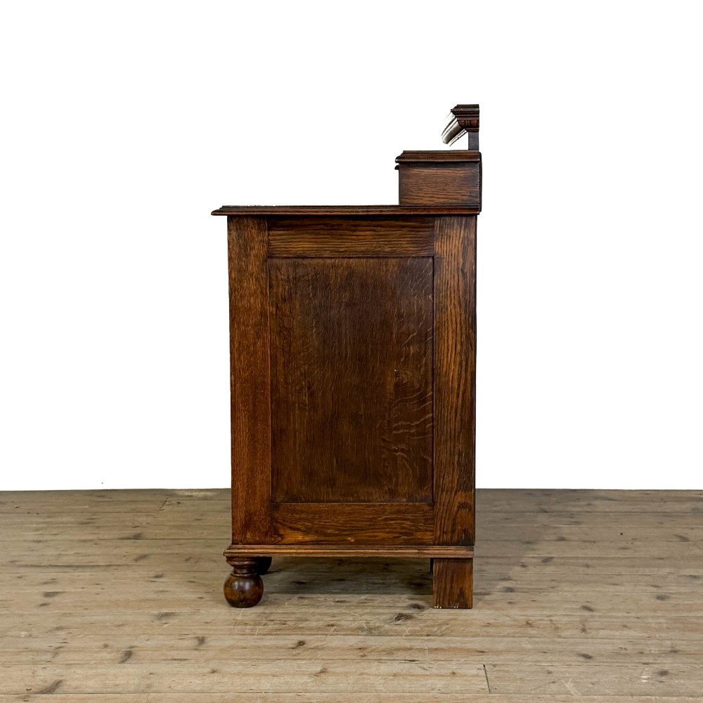 An Arts and Crafts solid oak kneehole desk featuring a raised top gallery shelf, central drawer with geometric moulding, drop handles, and side cupboards with circular gridded glass panels. View V