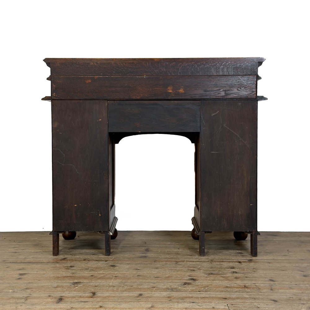 An Arts and Crafts solid oak kneehole desk featuring a raised top gallery shelf, central drawer with geometric moulding, drop handles, and side cupboards with circular gridded glass panels. View VI
