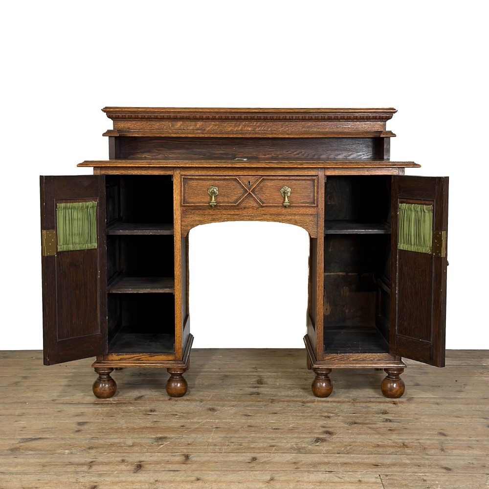 An Arts and Crafts solid oak kneehole desk featuring a raised top gallery shelf, central drawer with geometric moulding, drop handles, and side cupboards with circular gridded glass panels. View VII