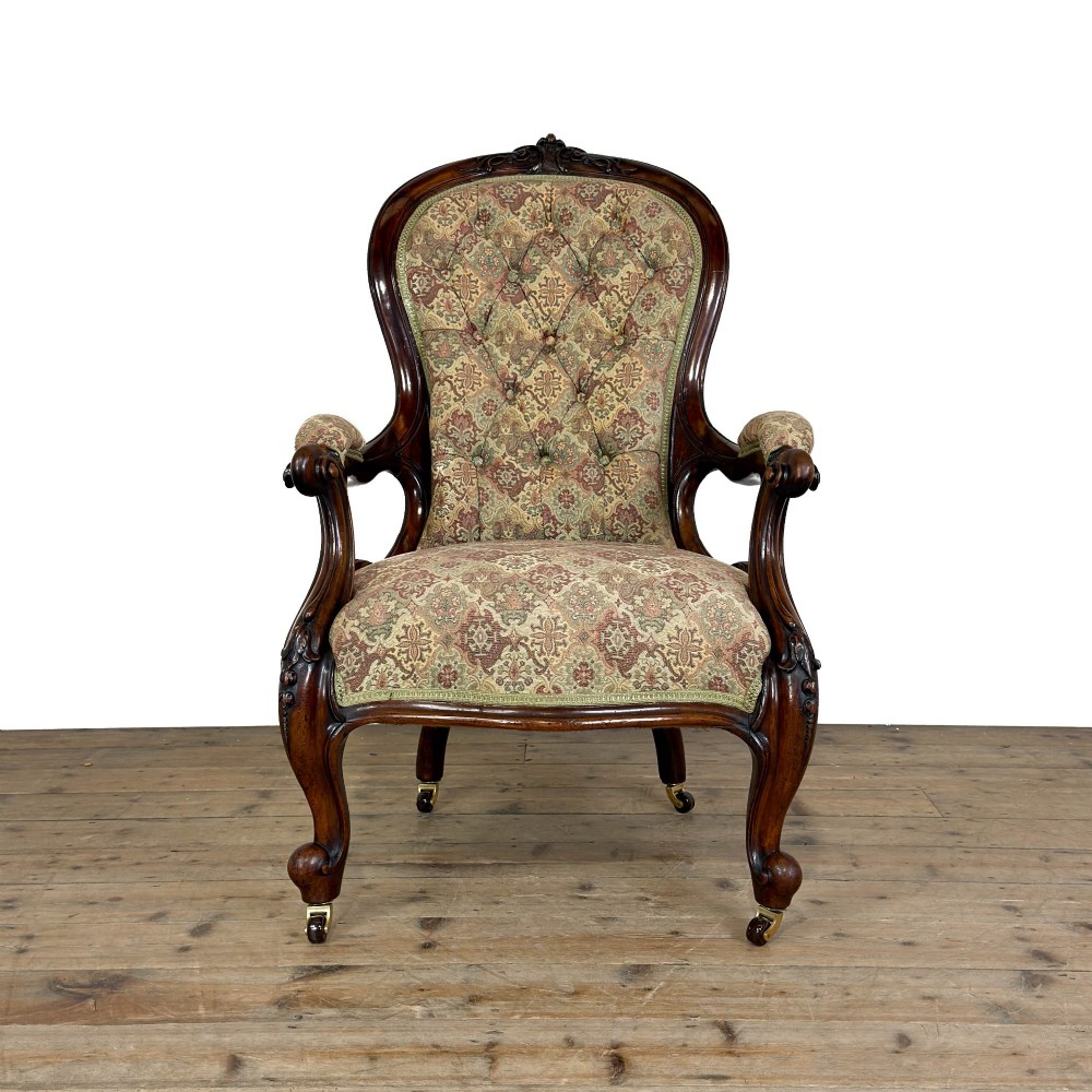 Victorian carved mahogany armchair with deep button-backed floral tapestry upholstery, curved armrests, and cabriole legs on brass castors.