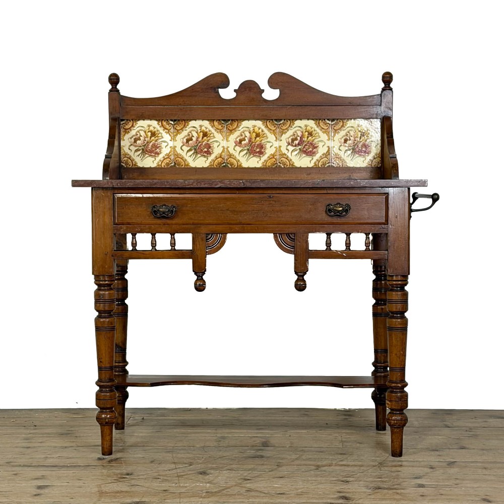 An Edwardian wooden washstand with a carved splashback housing floral glazed tiles, a single drawer with drop handles, turned legs, and an undertier shelf.