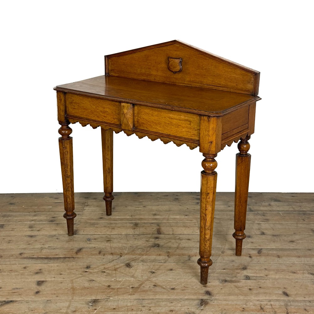 An antique warm brown oak hall table featuring a triangular gallery back with a central carved shield motif, a scalloped apron, and turned legs. View II