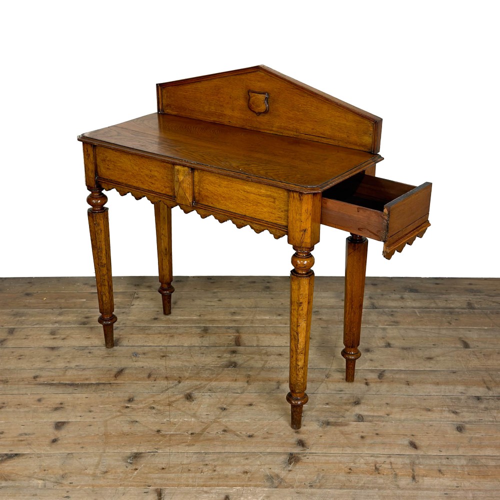 An antique warm brown oak hall table featuring a triangular gallery back with a central carved shield motif, a scalloped apron, and turned legs. View V