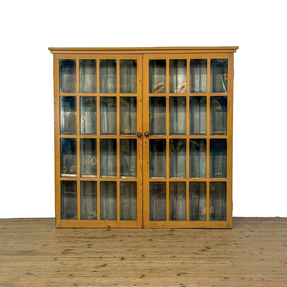 A large vintage pine cabinet featuring twin glazed doors with multiple glass panes and dark wooden knob handles.