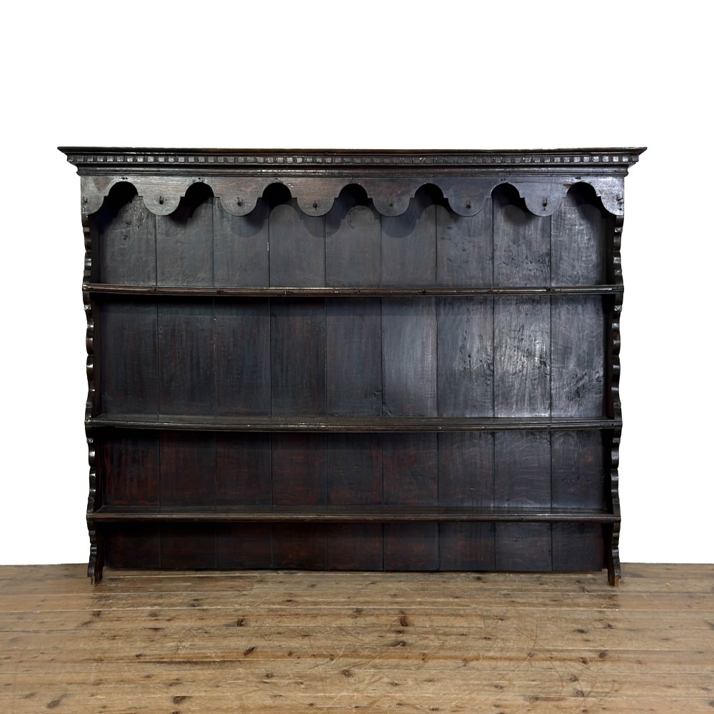 A dark oak wall-mounted plate rack featuring three open shelves, a scalloped top frieze with hooks, and dentil moulding along the upper cornice.
