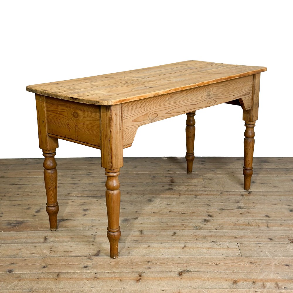 An antique stripped pine kitchen table featuring a plank top, a deep apron with corner cut-outs, and four turned legs.