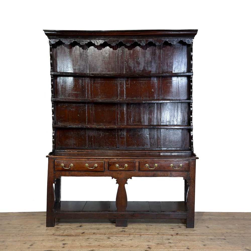 An eighteenth-century dark oak Welsh dresser featuring an open three-tier plate rack with a scalloped top frieze over a three-drawer base with brass handles and a lower potboard shelf.