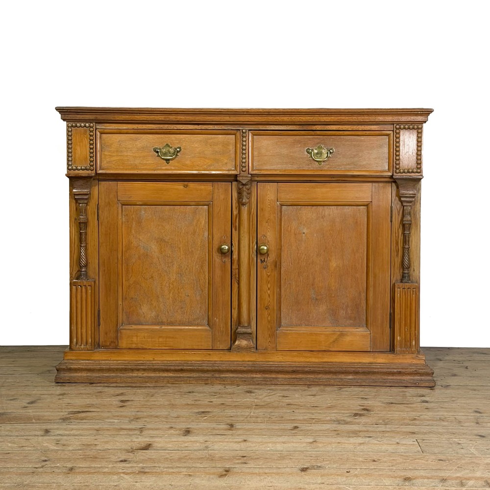 An antique golden brown oak sideboard featuring two drawers with brass handles over two panelled cupboard doors with carved side pilasters.