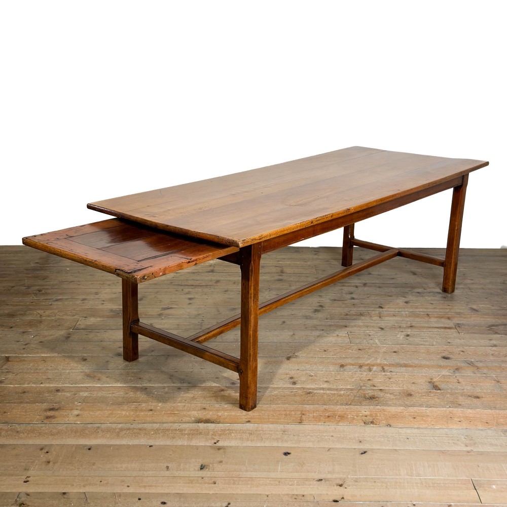 An antique fruitwood refectory dining table featuring a rectangular plank top, an extended slide-out leaf, square legs, and lower H-stretchers.
