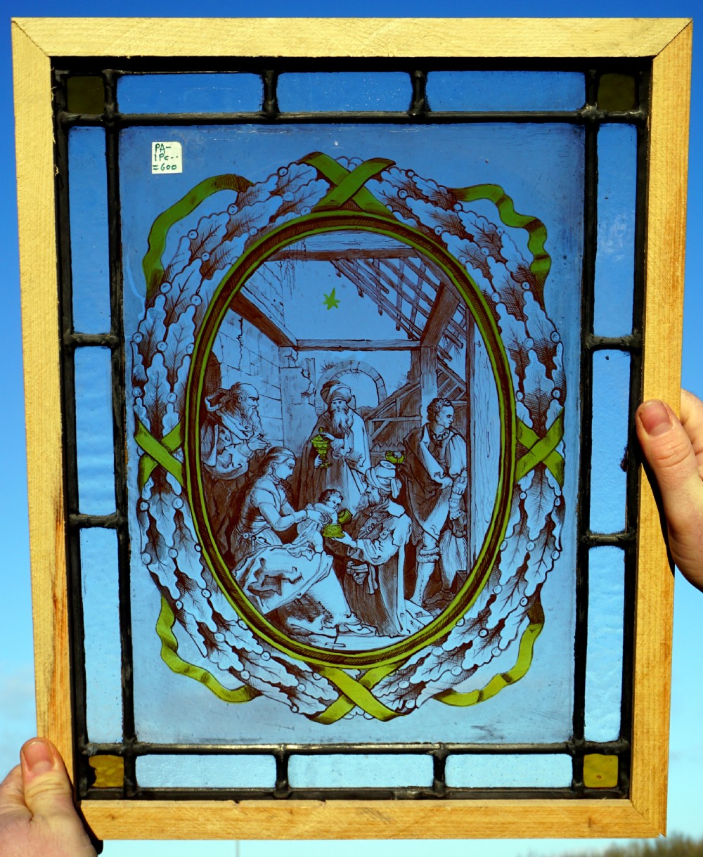 A rectangular framed stained glass panel featuring a central oval painting of a nativity scene enclosed by a leaf wreath and surrounded by cobalt blue glass.