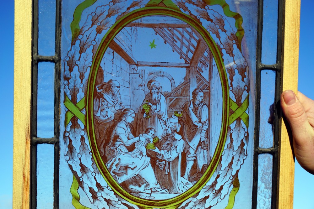 A rectangular framed stained glass panel featuring a central oval painting of a nativity scene enclosed by a leaf wreath and surrounded by cobalt blue glass. View II