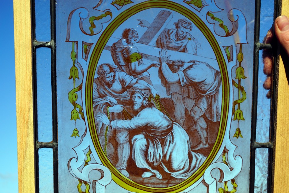A rectangular framed stained glass panel depicting a religious scene of Christ carrying the cross inside an ornate scrollwork oval border against a cobalt blue background. View II