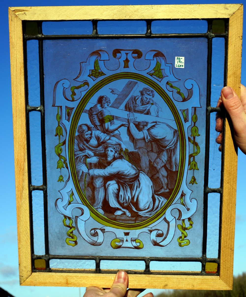 A rectangular framed stained glass panel depicting a religious scene of Christ carrying the cross inside an ornate scrollwork oval border against a cobalt blue background.