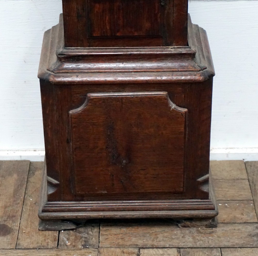 A dark oak granddaughter clock featuring a brass square dial with spandrels, carved cresting, an arched trunk door, and a moulded plinth base. View II