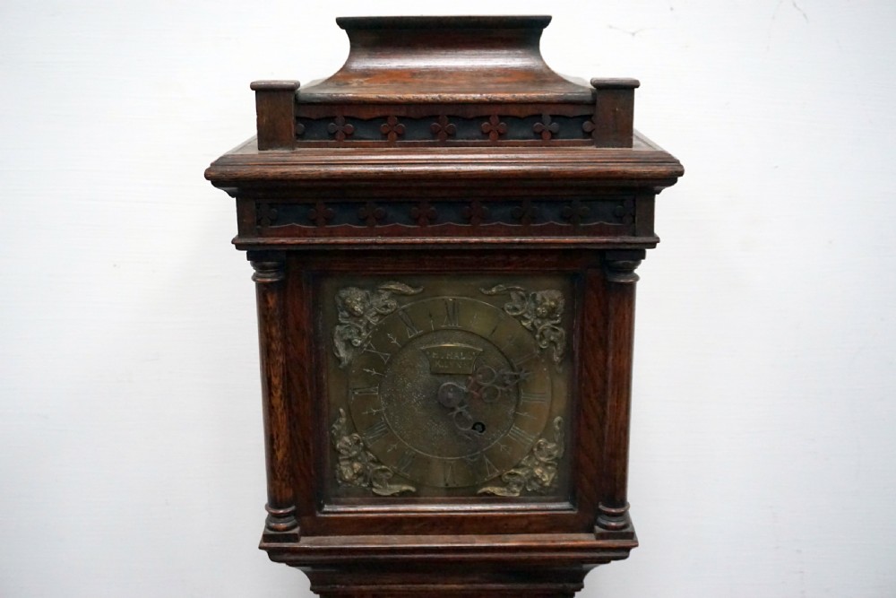A dark oak granddaughter clock featuring a brass square dial with spandrels, carved cresting, an arched trunk door, and a moulded plinth base. View IV