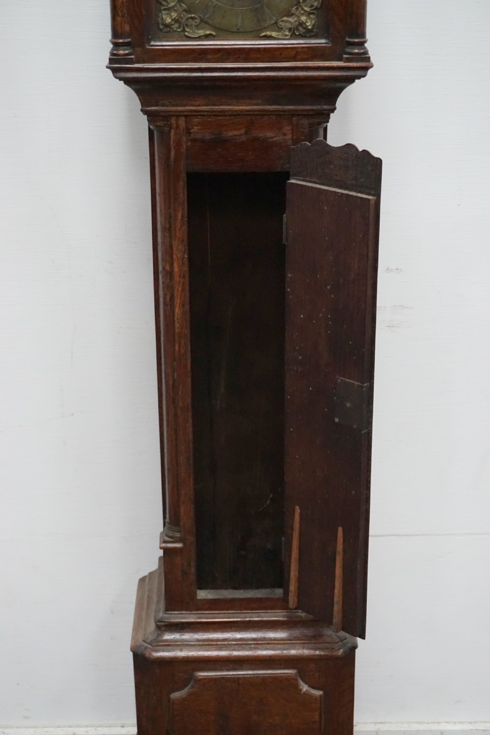 A dark oak granddaughter clock featuring a brass square dial with spandrels, carved cresting, an arched trunk door, and a moulded plinth base. View V