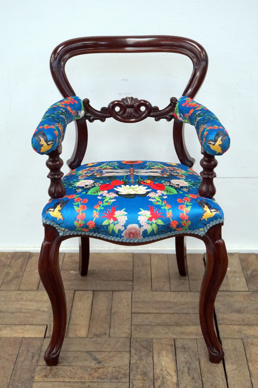 A Victorian mahogany armchair with carved scrollwork, padded armrests, cabriole legs, and vibrant blue upholstery featuring botanical floral and dragonfly patterns.
