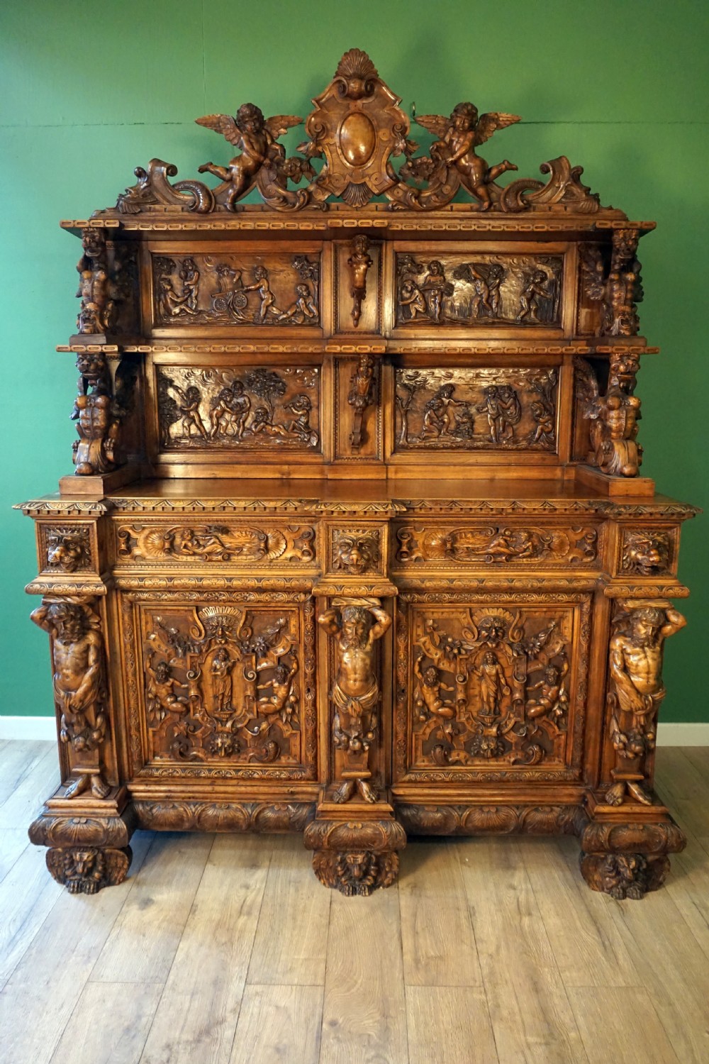 A two-tier carved wooden buffet featuring detailed relief panels of mythological scenes, carved putti, and supporting caryatid figures.
