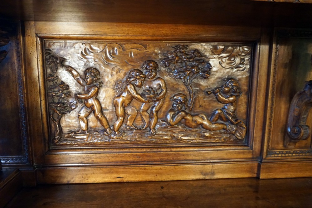 A two-tier carved wooden buffet featuring detailed relief panels of mythological scenes, carved putti, and supporting caryatid figures. View V