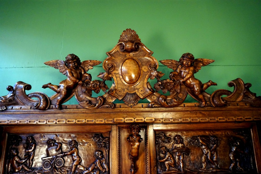 A two-tier carved wooden buffet featuring detailed relief panels of mythological scenes, carved putti, and supporting caryatid figures. View X
