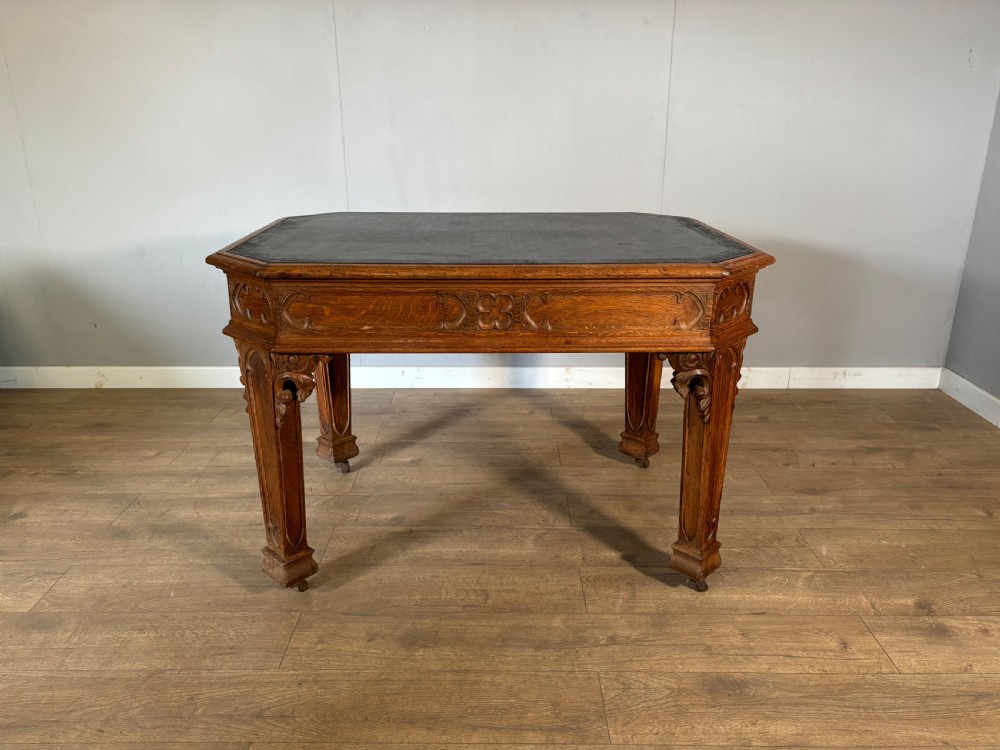A Victorian Gothic Revival oak writing table featuring a dark leather inset top, carved quatrefoil frieze details, and detailed corner legs on castors.