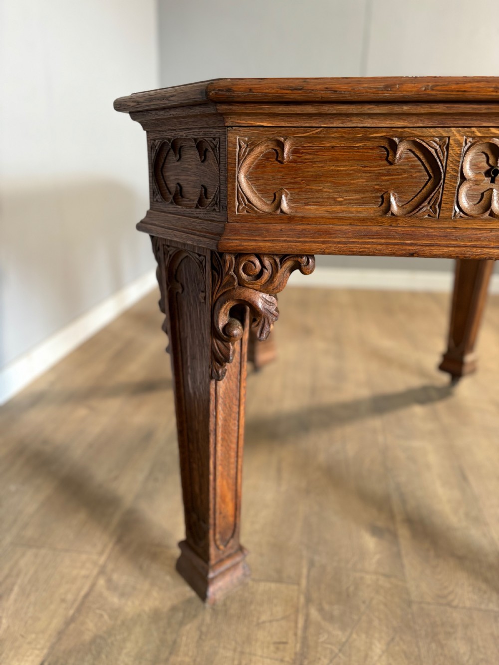 A Victorian Gothic Revival oak writing table featuring a dark leather inset top, carved quatrefoil frieze details, and detailed corner legs on castors. View V
